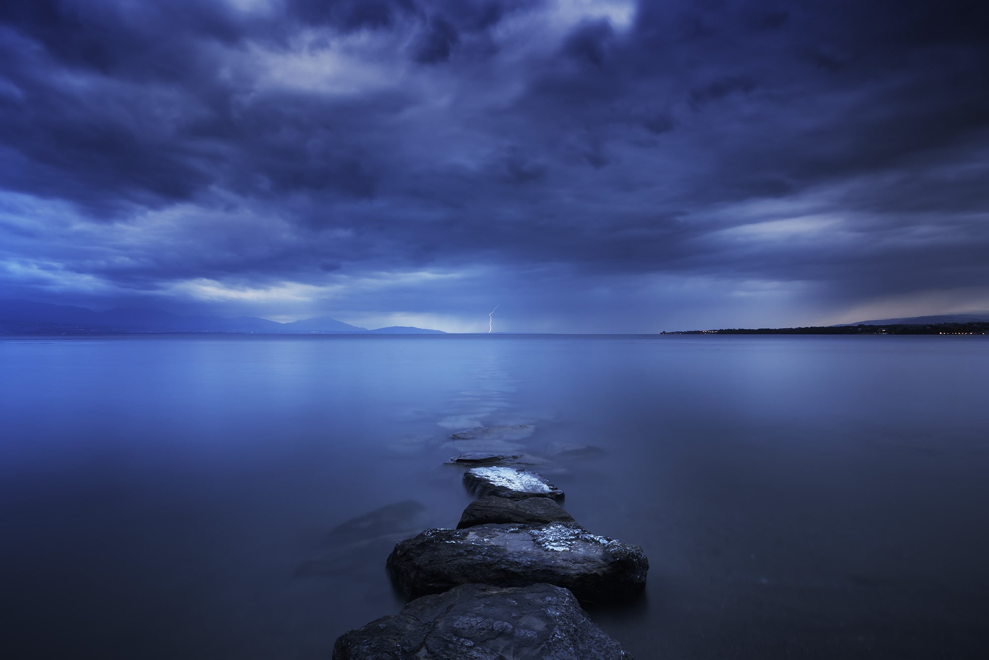 Landscape photography of Lake Geneva taken from Préverenges Beach during a dramatic summer storm in Switzerland, featuring lightning and a moody thunderstorm sky.