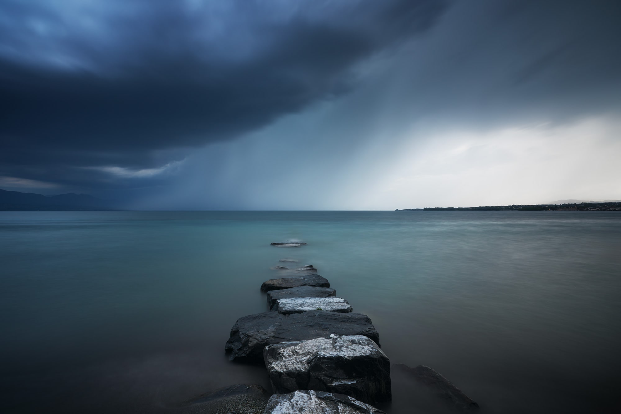 Stormy sky landscape photography showing a dramatic storm approaching over Lake Geneva, captured from Préverenges beach near Lausanne, Switzerland. Pink sunset light contrasts with the incoming clouds.
