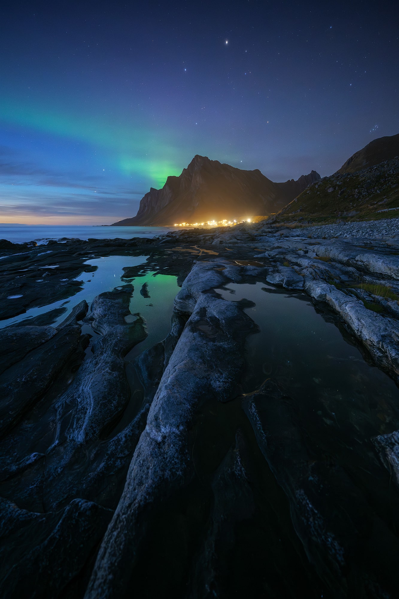 Wide-angle landscape photography of the northern lights during the blue hour in the Lofoten Islands, Norway. Green aurora borealis glows above the rugged Arctic coastline, with rocky foreground textures and reflections under a deep blue sky.