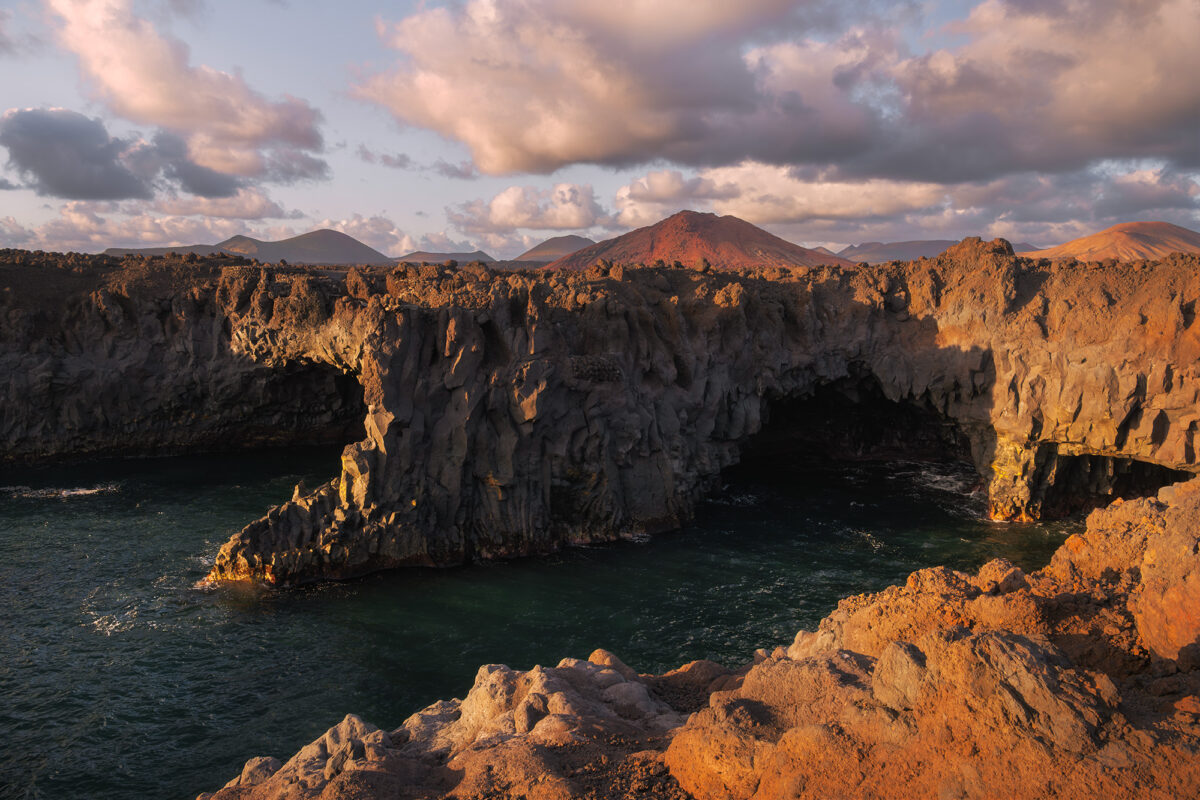 Landscape photography at sunset in Los Hervideros, Lanzarote, Canary Islands, with a volcano in the background and striking orange volcanic rocks along the rugged coastline.