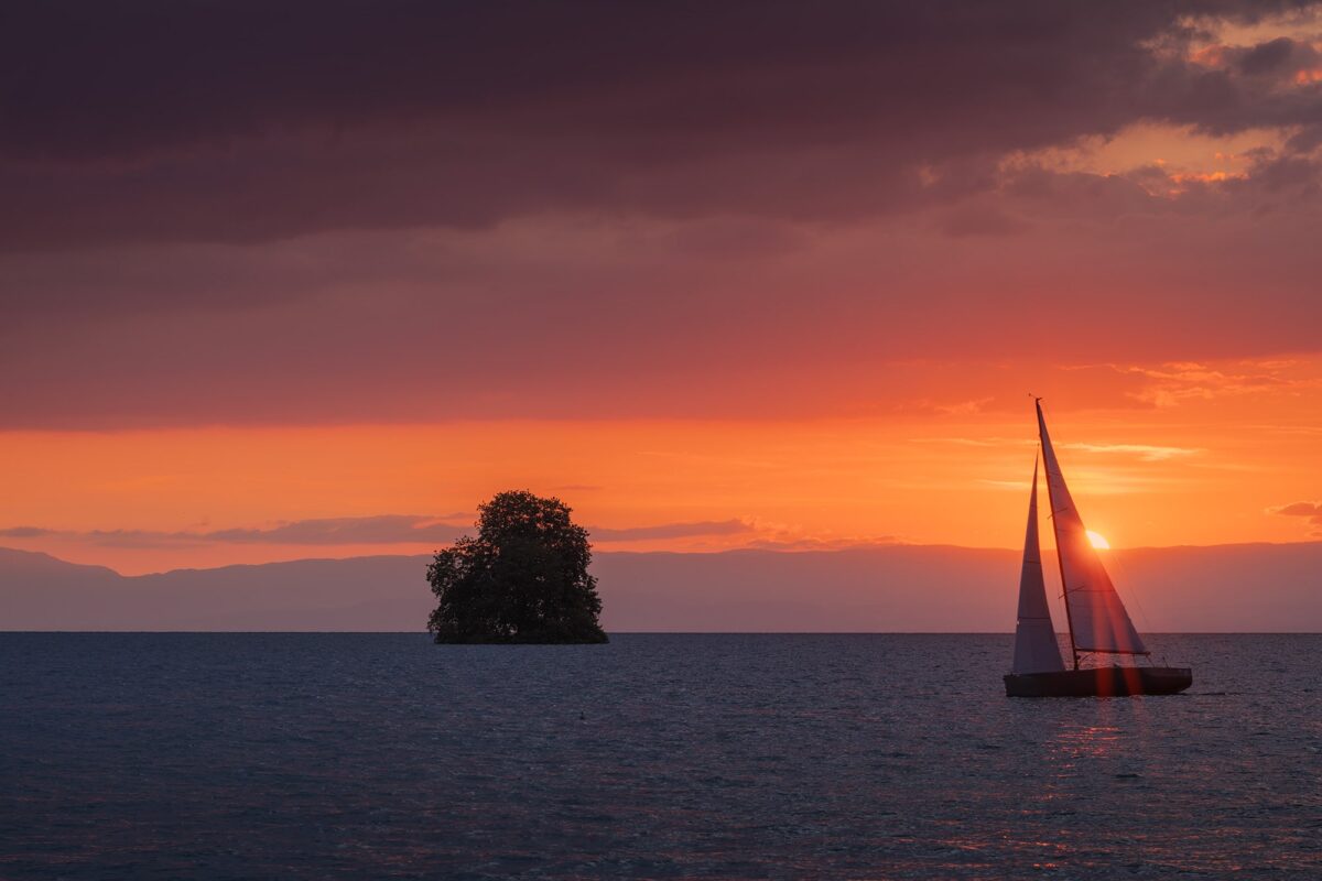 Sailboat sailing on Lake Geneva at sunset, photographed from Villeneuve, Switzerland — warm golden light reflecting on the water and mountains in the background.