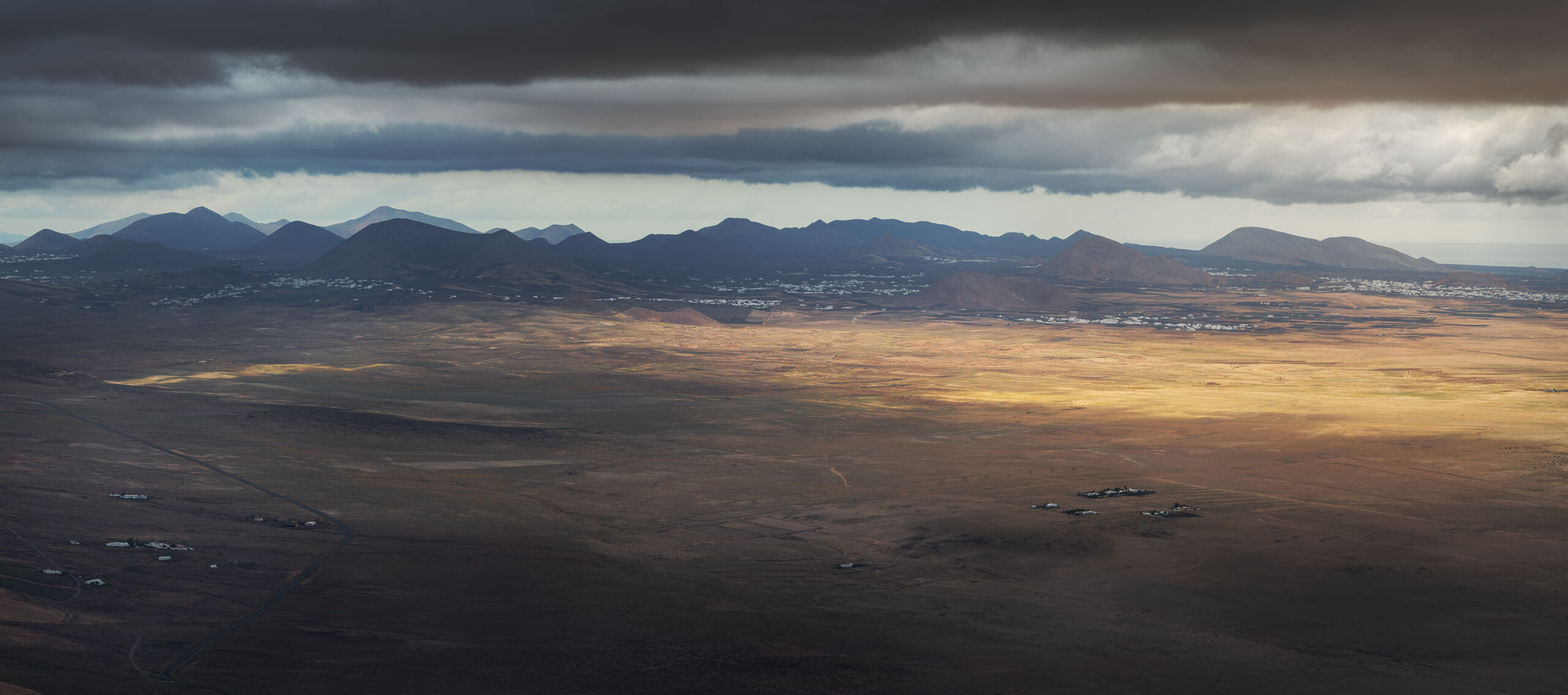 Panorama photography from Famara Viewpoint in Lanzarote, featuring the volcanic island’s dramatic peaks, cloudy and epic sky over the volcanoes, and sweeping Atlantic views.