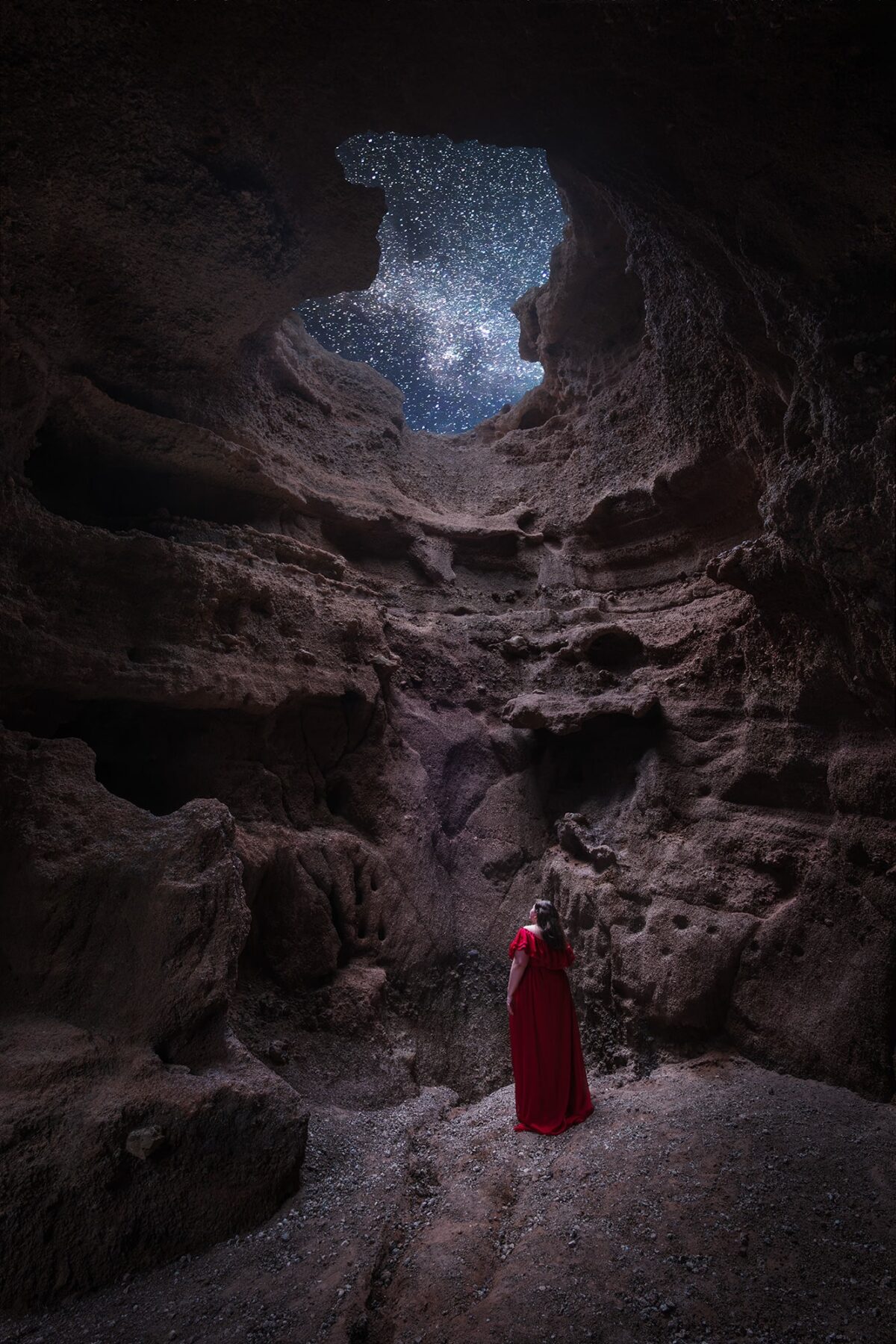 Self-portrait of a woman in a flowing red dress standing inside a cave in Lanzarote, Canary Islands, looking up at the stars through a large opening in the cave ceiling.