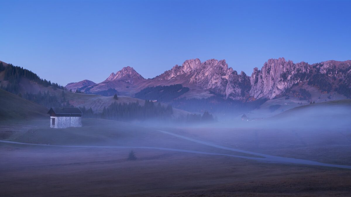 Landscape photography captured during the blue hour at Jaunpass in Simmental, Switzerland, showcasing the Gastlosen mountain range emerging above gentle fog and a solitary alpine cabin.