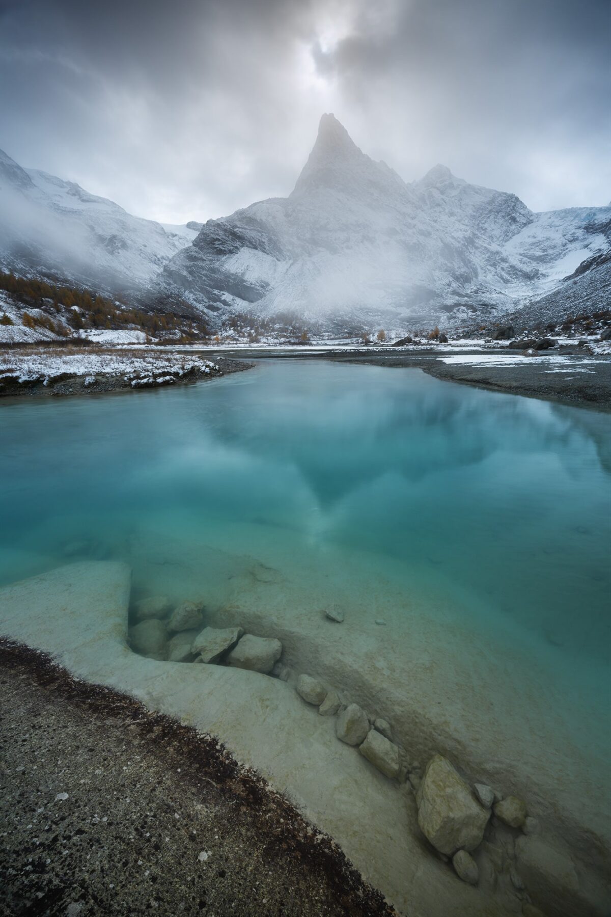 Moody autumn weather at Ferpècle in the Canton of Valais, with the mountain reflecting in the turquoise river fed by the glacier. Light fog and clouds linger after the first snowfall of October, creating a dramatic alpine atmosphere.