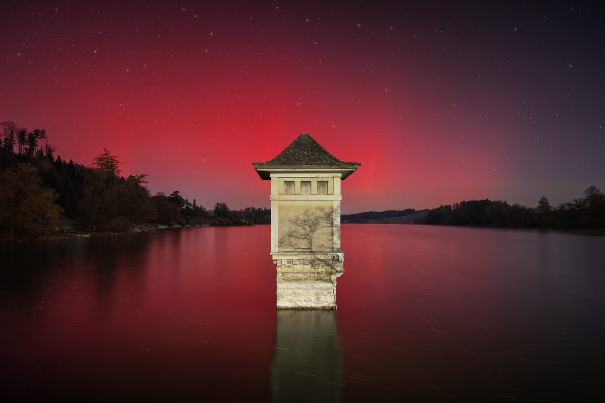Red northern lights in Switzerland captured on November 12, 2025, by Jennifer Esseiva at Lac de Bret in the Canton of Vaud, near Chexbres. The aurora glows above the calm lake, reflecting vibrant crimson hues in the night sky.