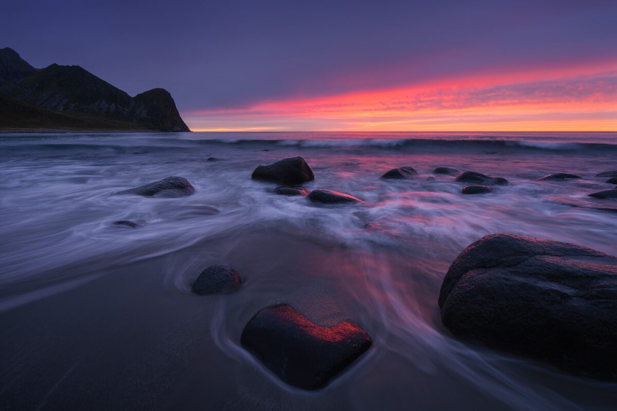 Long-exposure landscape photography of a red sunset at Unstad Beach in the Lofoten, Norway, featuring a heart-shaped rock in the foreground under dramatic glowing skies.