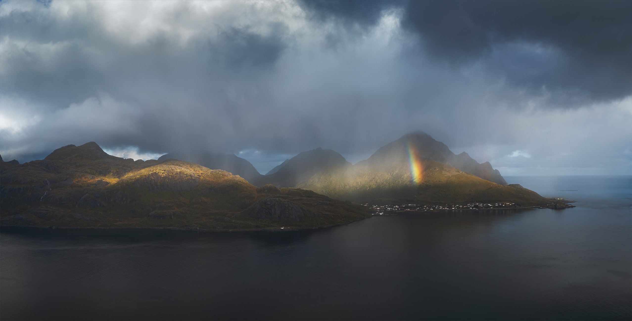 Drone photography of the Lofoten Islands, Norway, captured on a moody day with dramatic clouds and a vivid rainbow illuminating the mountains above a calm Arctic fjord.
