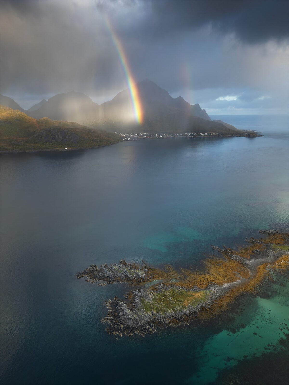Drone photography of the Lofoten Islands, Norway, captured on a moody day with dramatic clouds and a vivid rainbow illuminating the mountains above a calm Arctic fjord.