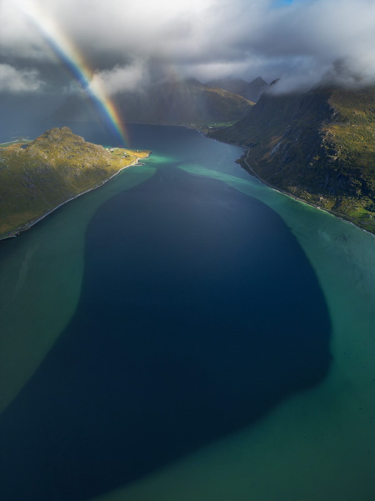 Drone landscape photography of Flakstadpollen in the Lofoten Islands, Norway, captured on a moody day with a vivid rainbow illuminating the mountains and turquoise fjord below.