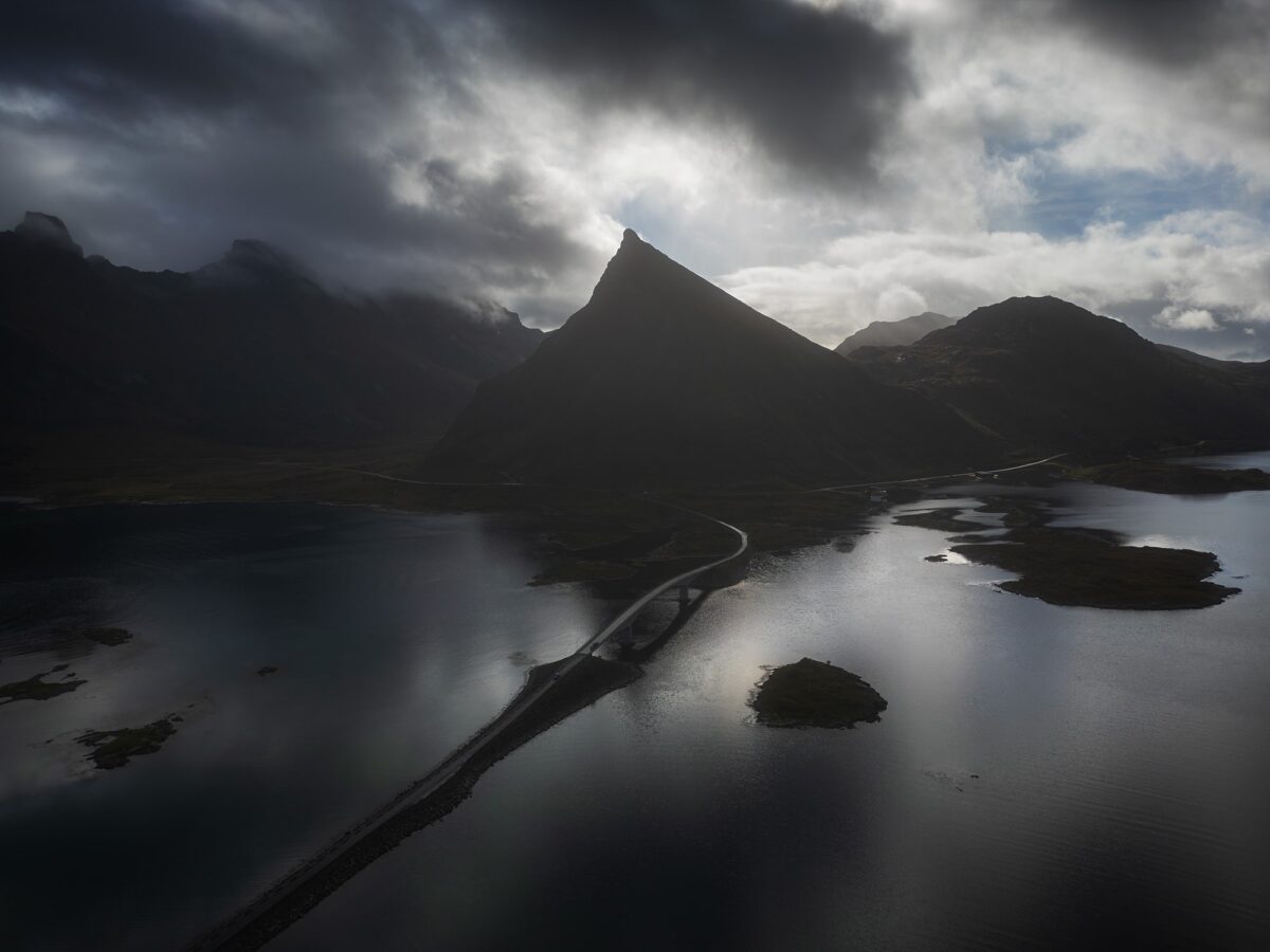Drone landscape photography of the Fredvang Bridges in the Lofoten Islands, Norway, captured on a moody day with dramatic clouds, dark tones, and reflections across the surrounding fjords.