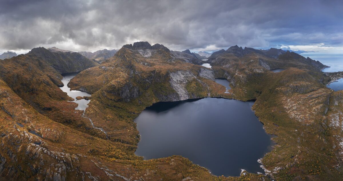 Panoramic drone landscape photography of the Tindstinden hike trailhead near Reine in the Lofoten Islands, Norway, captured in September with autumn colours and alpine lakes surrounded by rugged mountains.