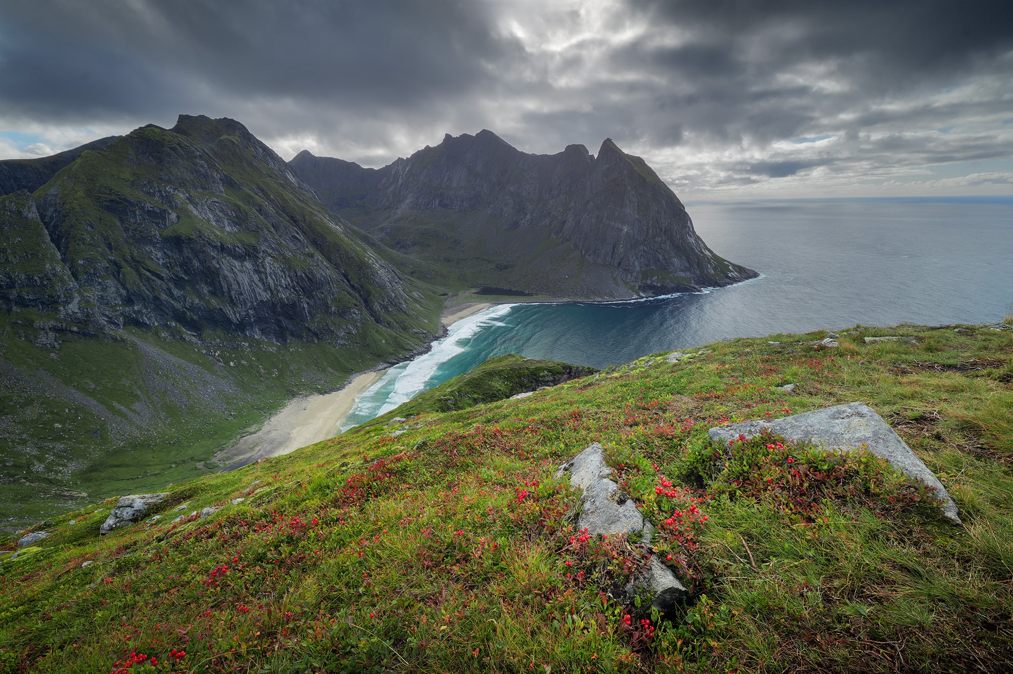 Landscape photography of Kvalvika Beach seen from the Ryten viewpoint in the Lofoten Islands, Norway, with dramatic cloud coverage and a wild, rugged Arctic landscape.