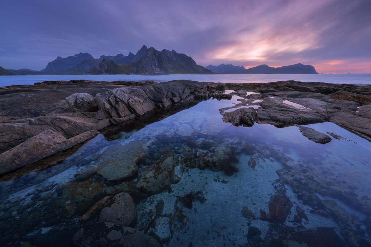 Landscape photography at sunset near Vareid in the Lofoten Islands, Norway, captured with a Nikon Z8, showing a reflective tidal pool in the rocky foreground and dramatic mountains under pastel evening skies.