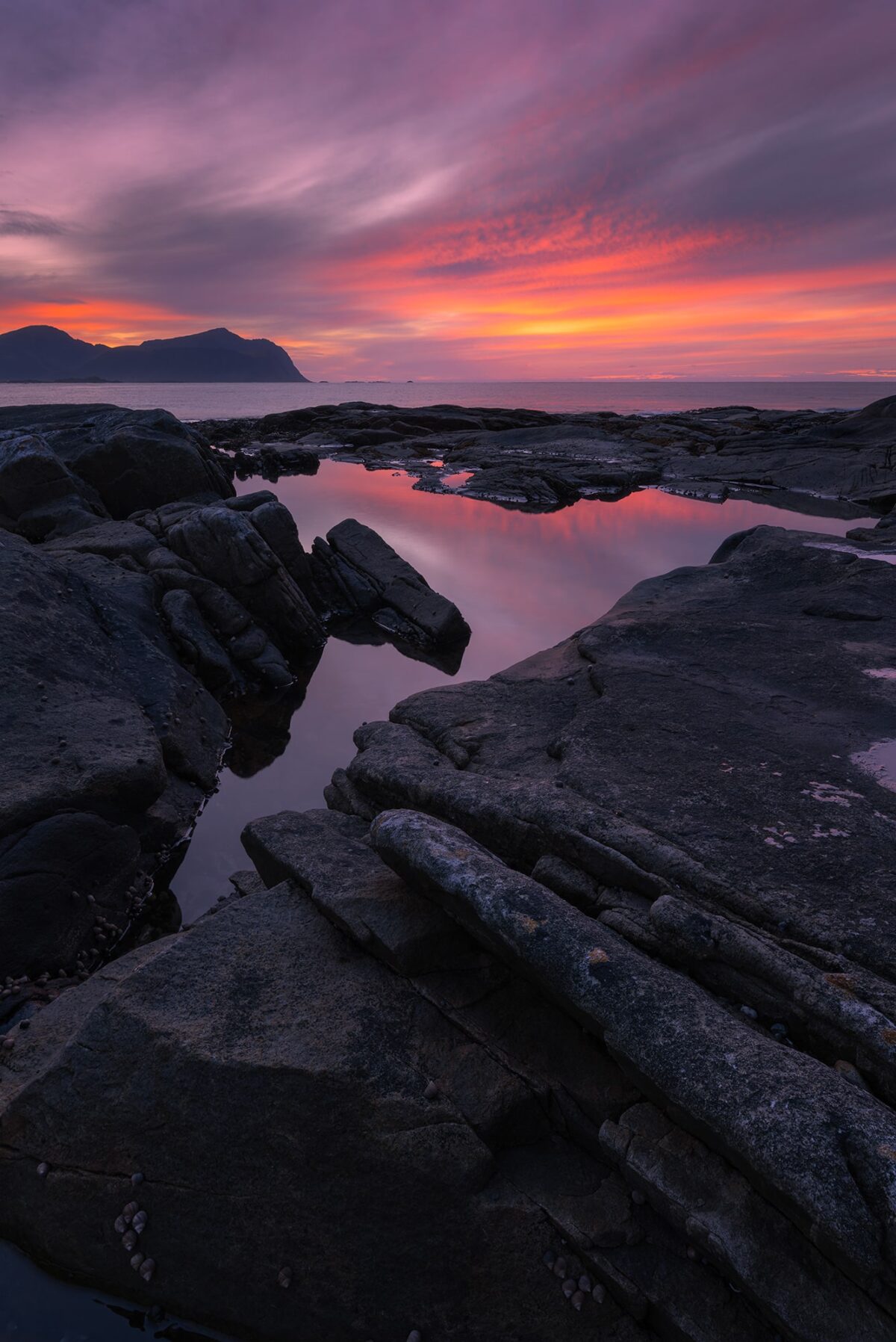 Landscape photography at sunset near Vareid in the Lofoten Islands, Norway, featuring a reflective tidal pool framed by dark volcanic rocks under a dramatic red and pink sky.