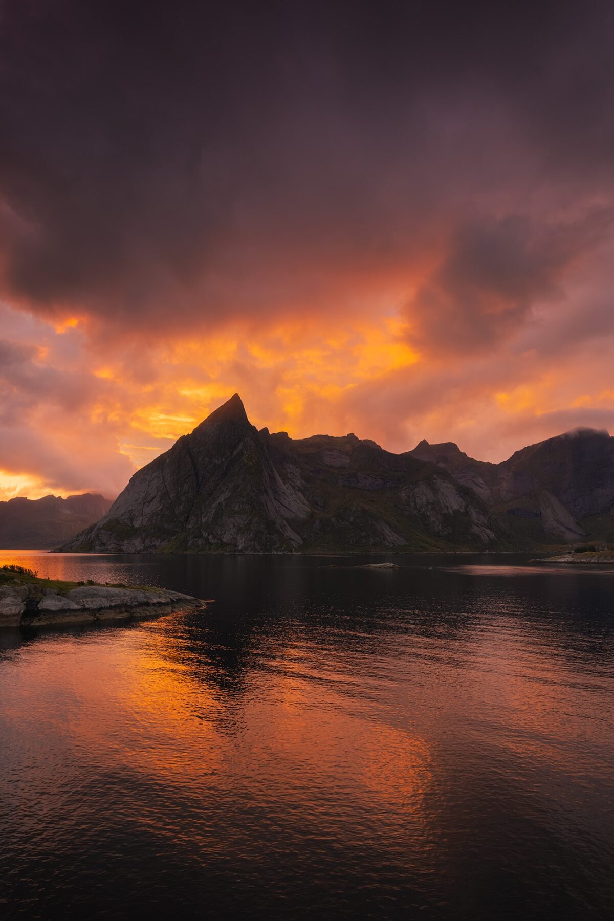 Landscape photography of Hamnøy in the Lofoten Islands, Norway, captured at sunset with dramatic orange clouds and a fiery sky reflecting on the calm water of the fjord beneath rugged mountain peaks.