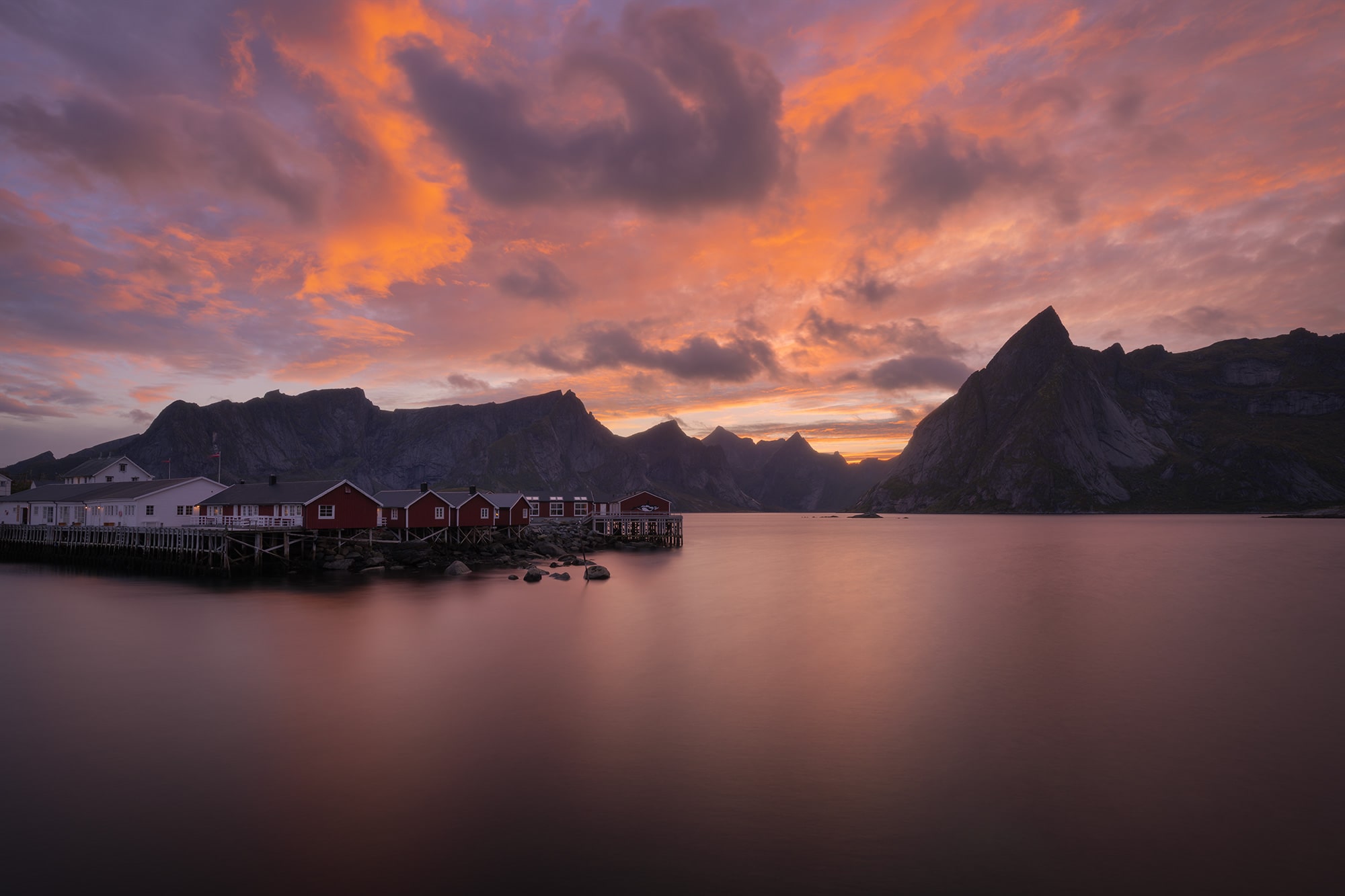 Landscape photography of Hamnøy fishing village in the Lofoten Islands, Norway, captured at sunset with glowing orange clouds above the iconic red rorbuer reflected in the calm Arctic water.