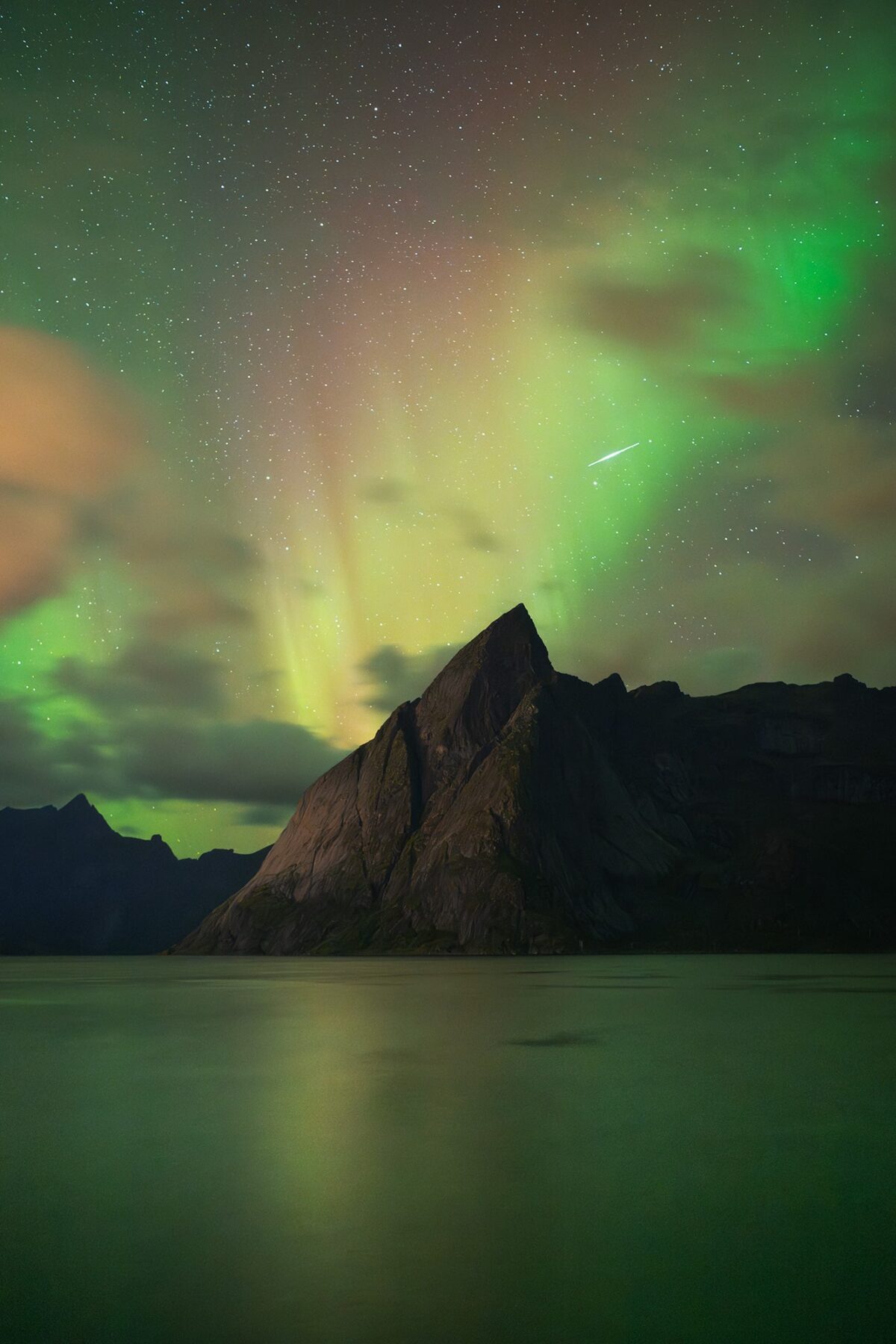 Landscape photography of fall northern lights above Reinefjorden in the Lofoten Islands, Norway, with a shooting star crossing the Arctic sky above the calm fjord and surrounding mountains.