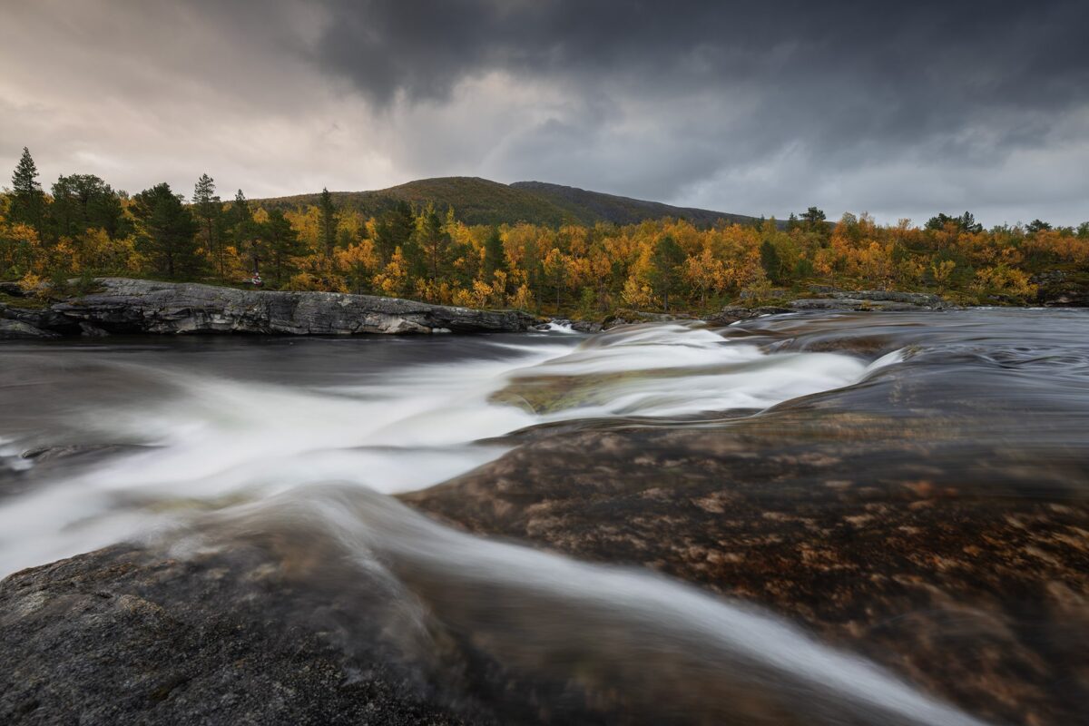 Landscape photography of Valnesfossen waterfall near the Bodø region in Norway, captured in September during a moody autumn sunset, with fall forest colours surrounding the cascading water.