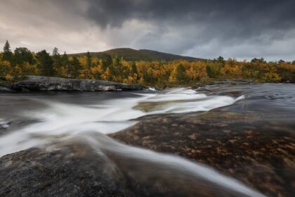 Landscape photography of Valnesfossen waterfall near the Bodø region in Norway, captured in September during a moody autumn sunset, with fall forest colours surrounding the cascading water.
