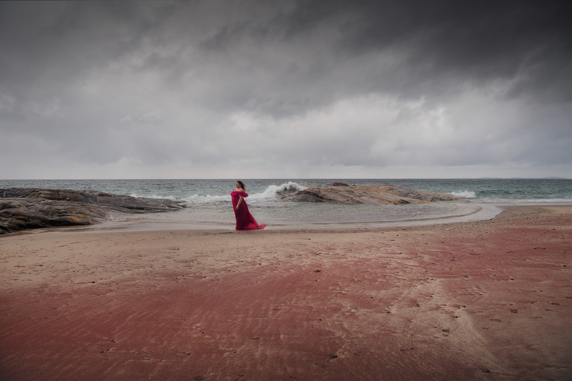 Self-portrait landscape photography of Jennifer Esseiva wearing a red dress on the red sand of Mjelle Beach near Bodø, Norway, captured in soft coastal light along the Arctic shoreline.