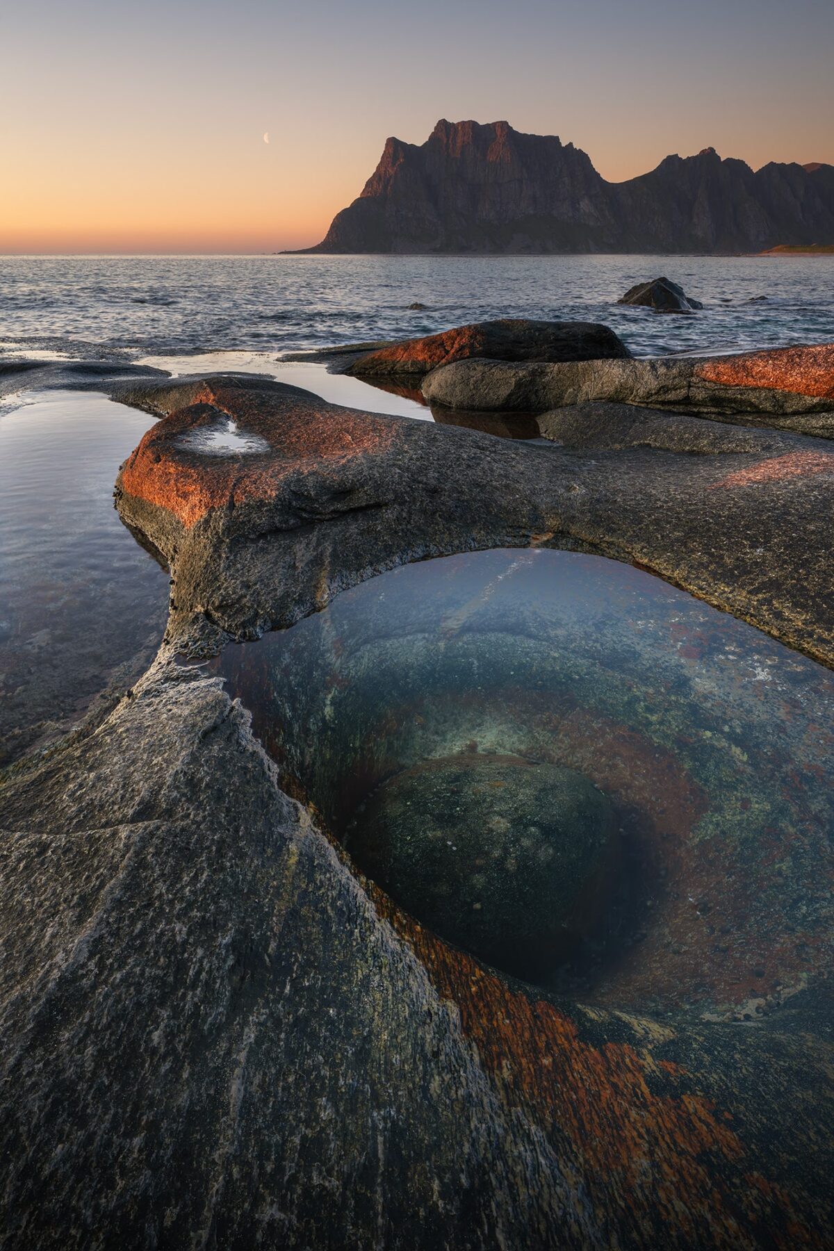 Vertical landscape photography of the Dragon Eye rock formation at Uttakleiv Beach in the Lofoten Islands, Norway, captured at sunset with a moonrise above the dramatic Arctic coastline.