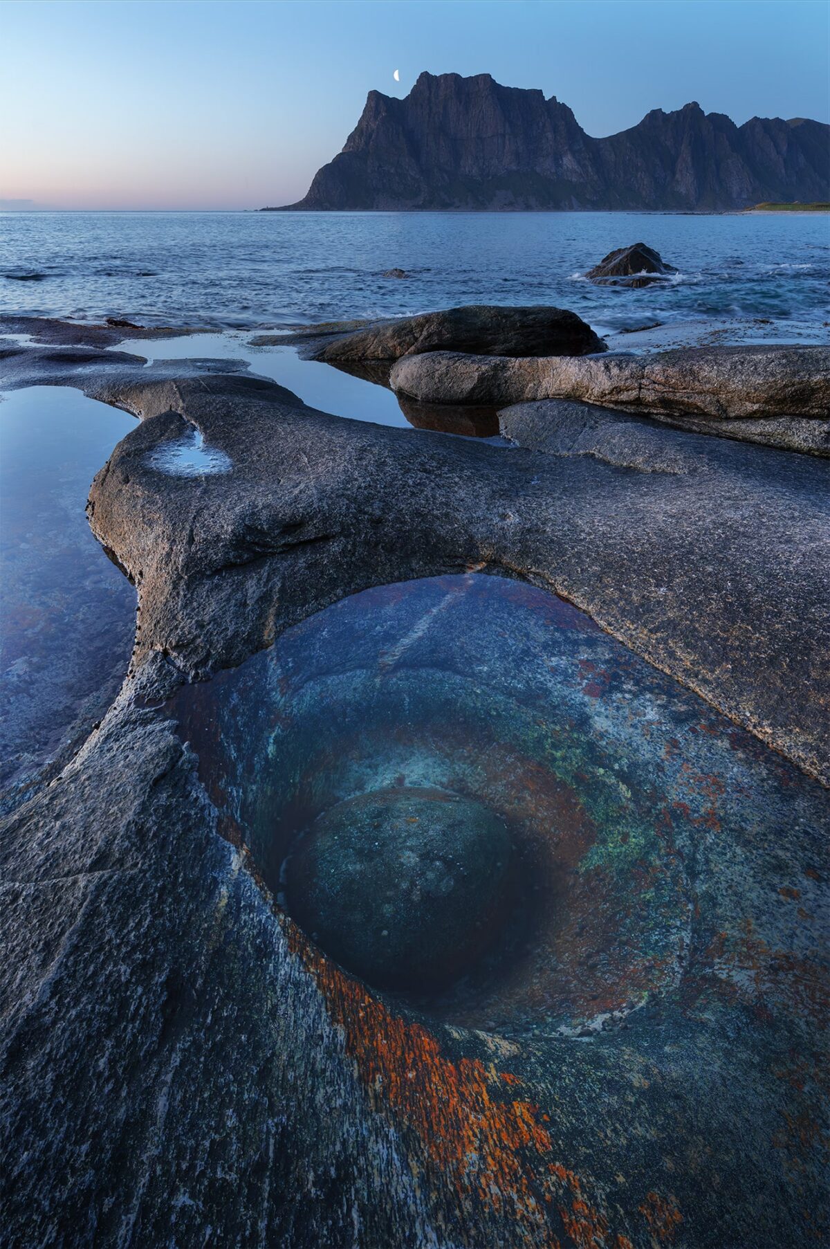 Vertical landscape photography of the Dragon Eye rock formation at Uttakleiv Beach in the Lofoten Islands, Norway, captured during the blue hour with the moon rising above the Arctic coastline.
