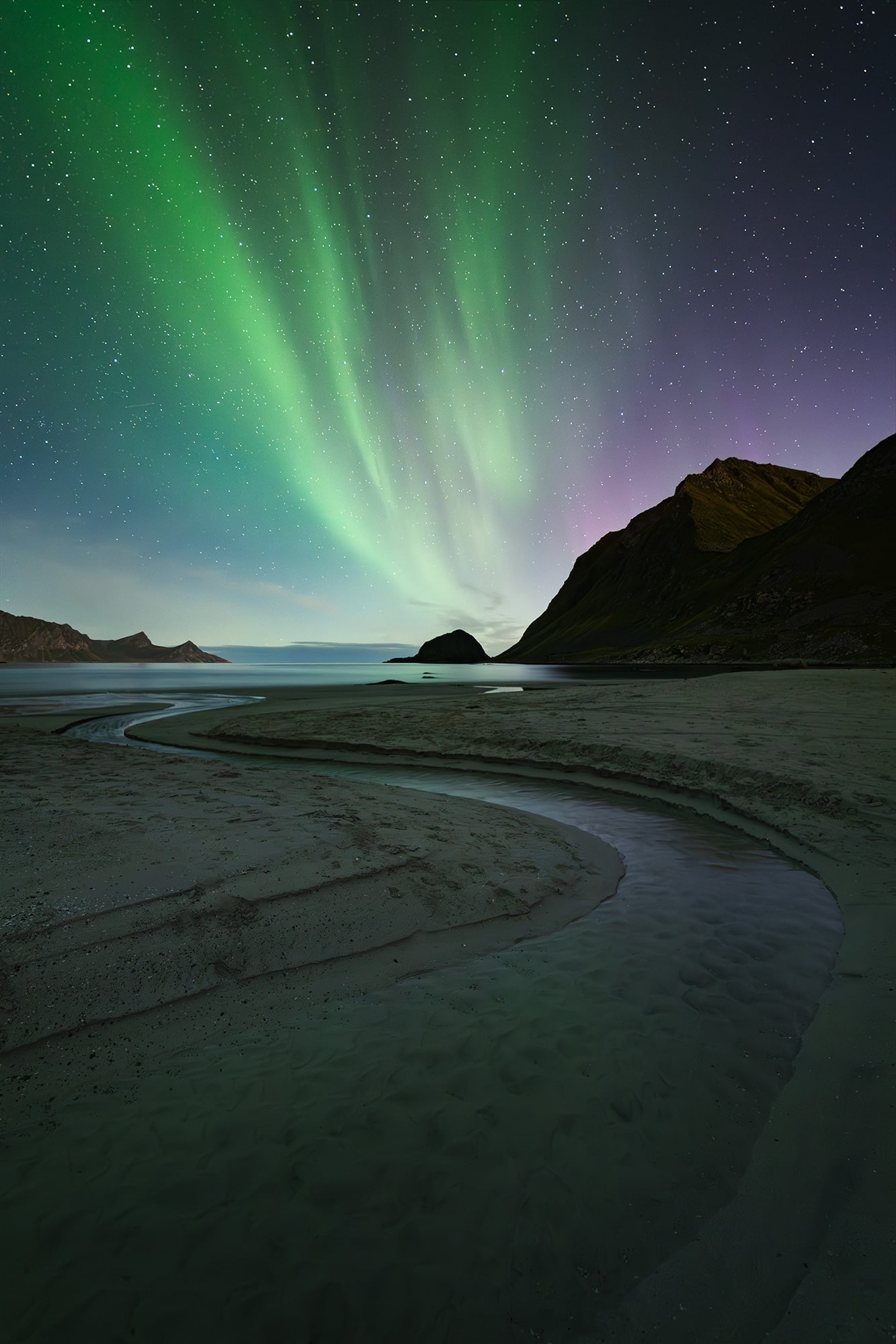 Landscape photography of Haukland Beach in the Lofoten Islands, Norway, captured at night under the northern lights, with an S-shaped river flowing into the sea in the foreground.