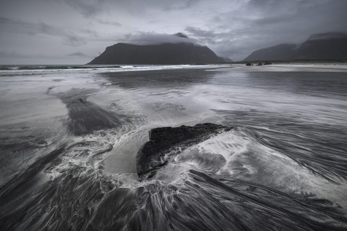 Landscape photography of Skagsanden Beach in the Lofoten Islands, Norway, captured in September during a moody autumn day, with dramatic cloud coverage and unique black sand patterns along the shoreline.