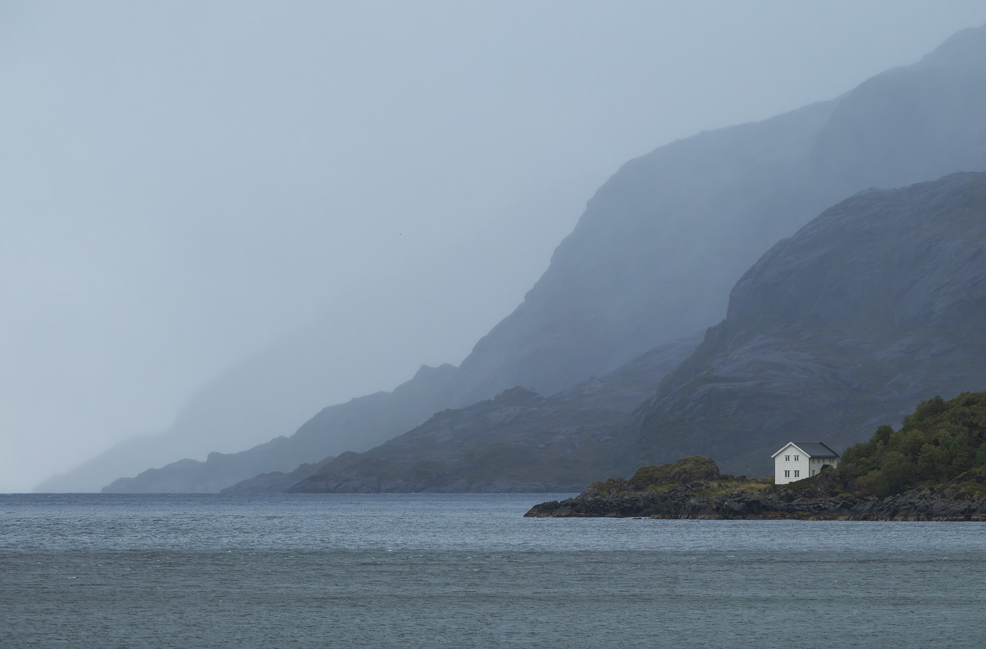 Landscape photography of a lonely white cabin on the coast of the Lofoten Islands, Norway, captured in moody weather with dramatic light and a quiet Arctic atmosphere.