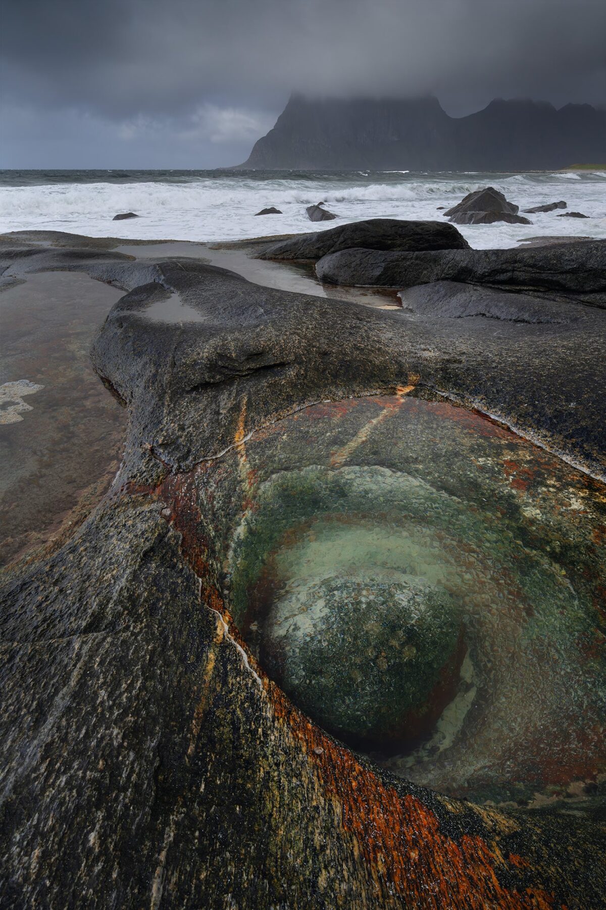 Vertical landscape photography of the Dragon Eye rock formation at Uttakleiv Beach in the Lofoten Islands, Norway, captured during a moody and windy blue hour with crashing waves, rain, and the moon rising above the Arctic coastline.