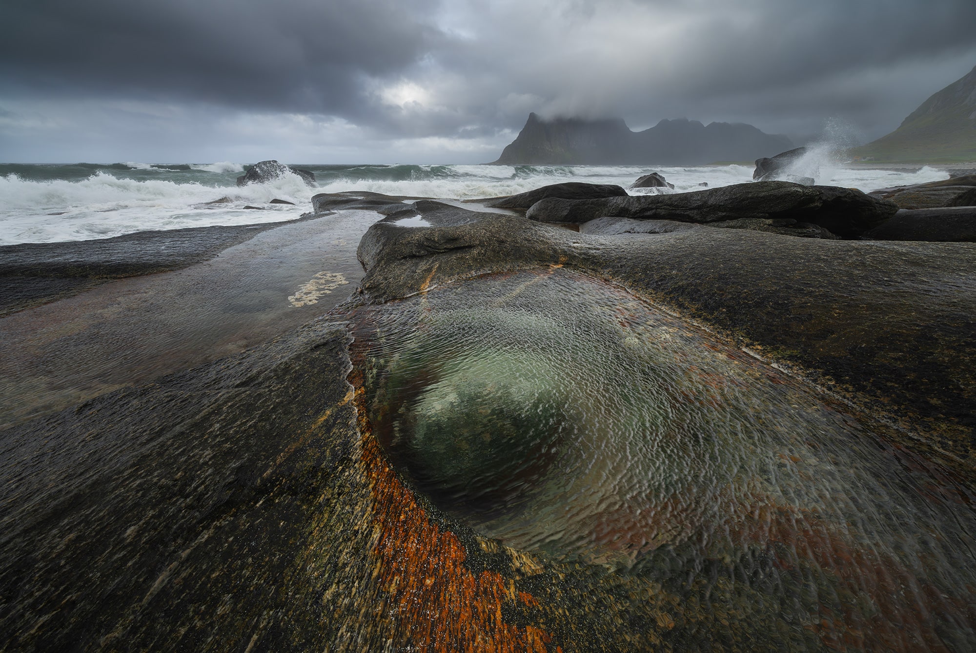 Horizontal landscape photography of the Dragon Eye rock formation at Uttakleiv Beach in the Lofoten Islands, Norway, captured during a moody and windy blue hour with crashing waves, rain, and the moon rising above the Arctic coastline.