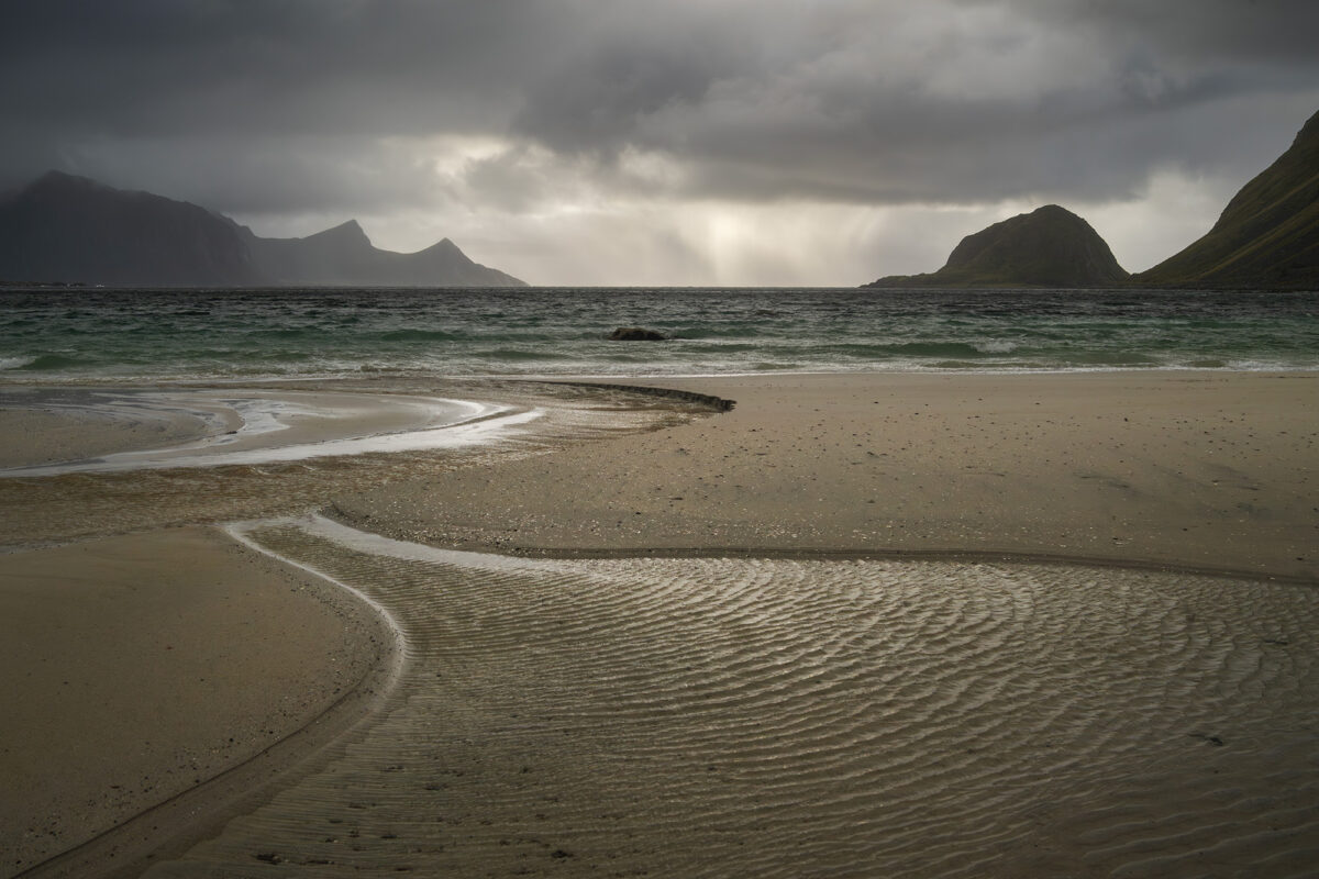 Landscape photography of Haukland Beach in the Lofoten Islands, Norway, captured during a stormy autumn day. An S-shaped stream flows across the sand into the sea, with dark clouds and dramatic light over the Arctic coastline.