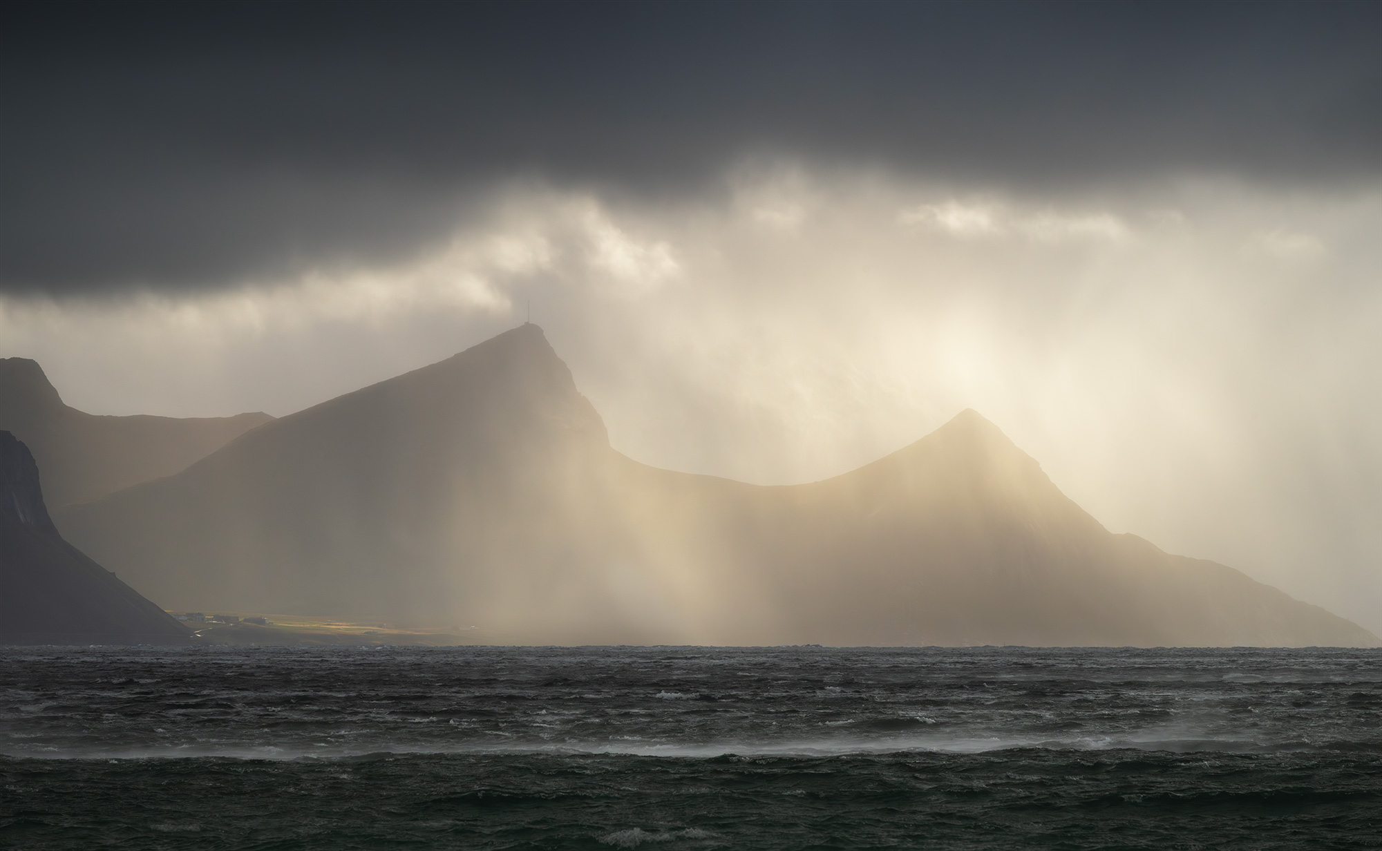 Landscape photography of the Lofoten Islands coastline, Norway, captured during a stormy day with dramatic rays of light breaking through dark clouds and illuminating the mountains above the Arctic sea.