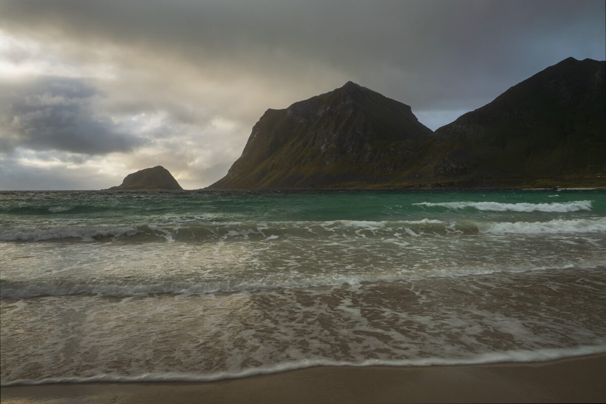 Landscape photography of Vik Strand in the Lofoten Islands, Norway, captured during a moody day with soft light, rolling waves, and dark mountains rising above the Arctic coastline.