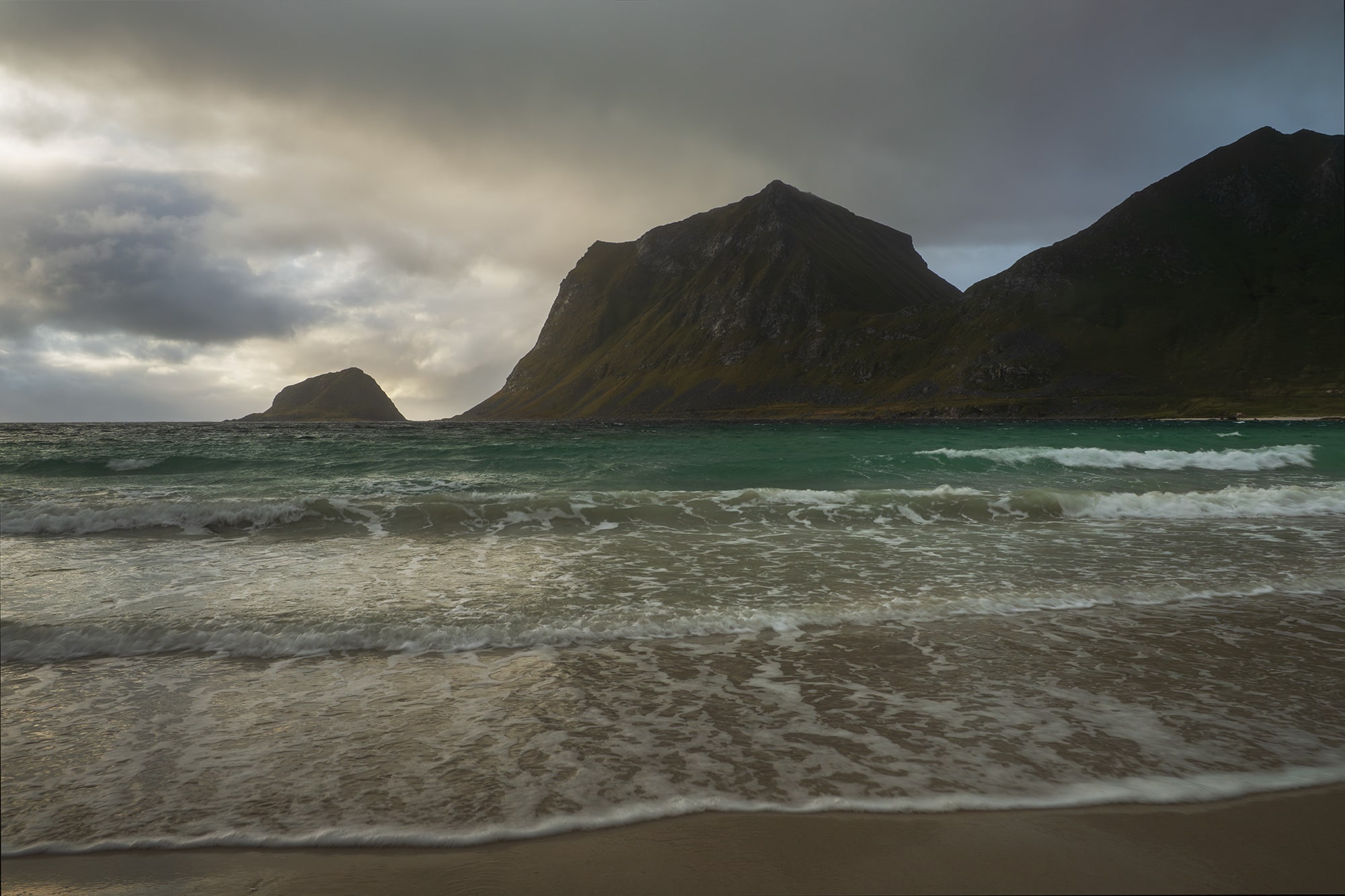 Landscape photography of Vik Strand in the Lofoten Islands, Norway, captured during a moody day with soft light, rolling waves, and dark mountains rising above the Arctic coastline.