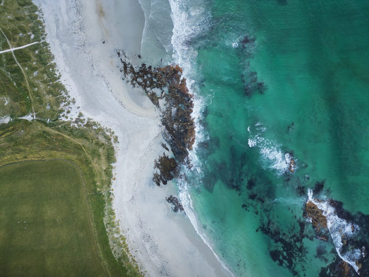 Aerial view of turquoise sea and white sand shoreline at Skagsanden Beach, Lofoten Islands, Norway, with green fields and rocky coast.