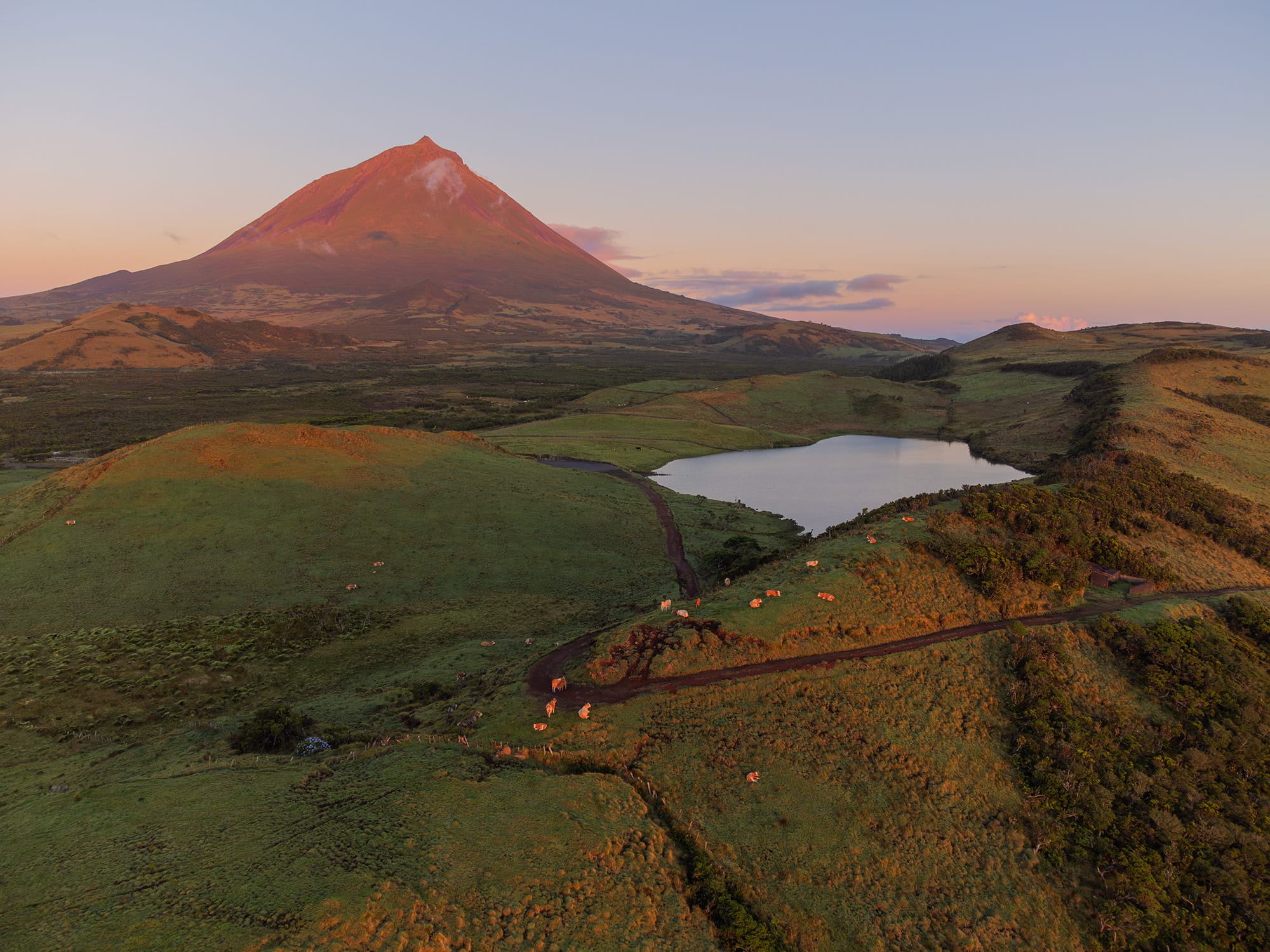 Aerial landscape photo of Lagoa do Capitão and Mount Pico at dawn, captured from a drone. The scene showcases pastel sunrise tones, still water and the volcanic silhouette.