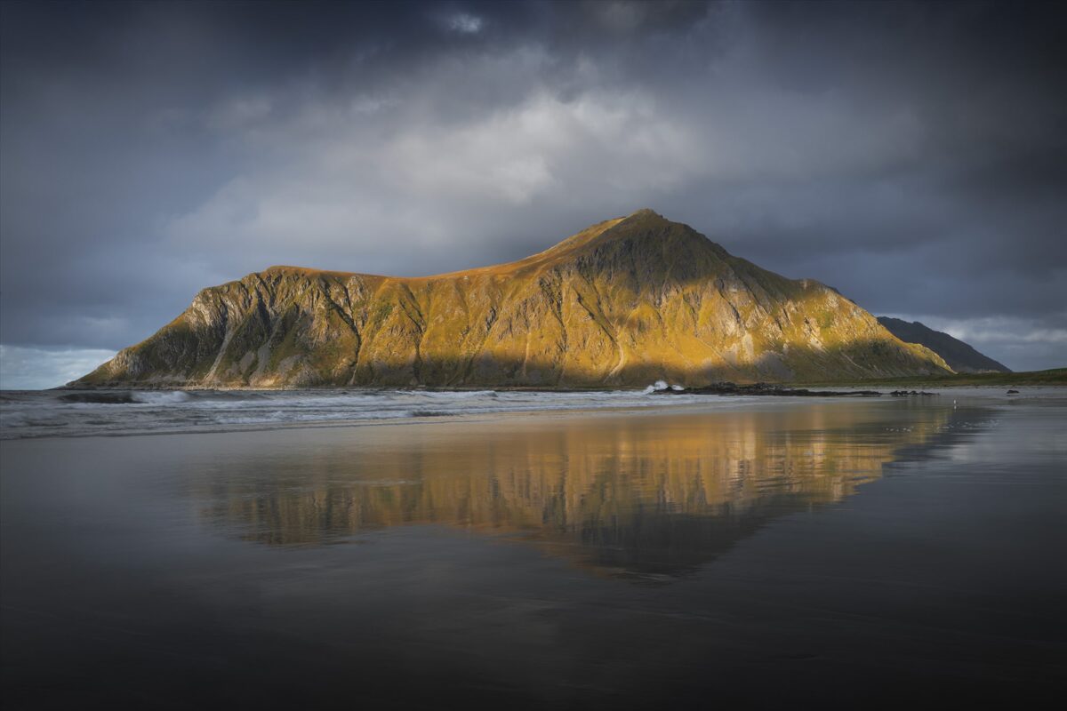 Golden sunlight hitting mountain at Skagsanden Beach, Lofoten Islands, Norway, with dramatic sky and reflection on wet sand during autumn.