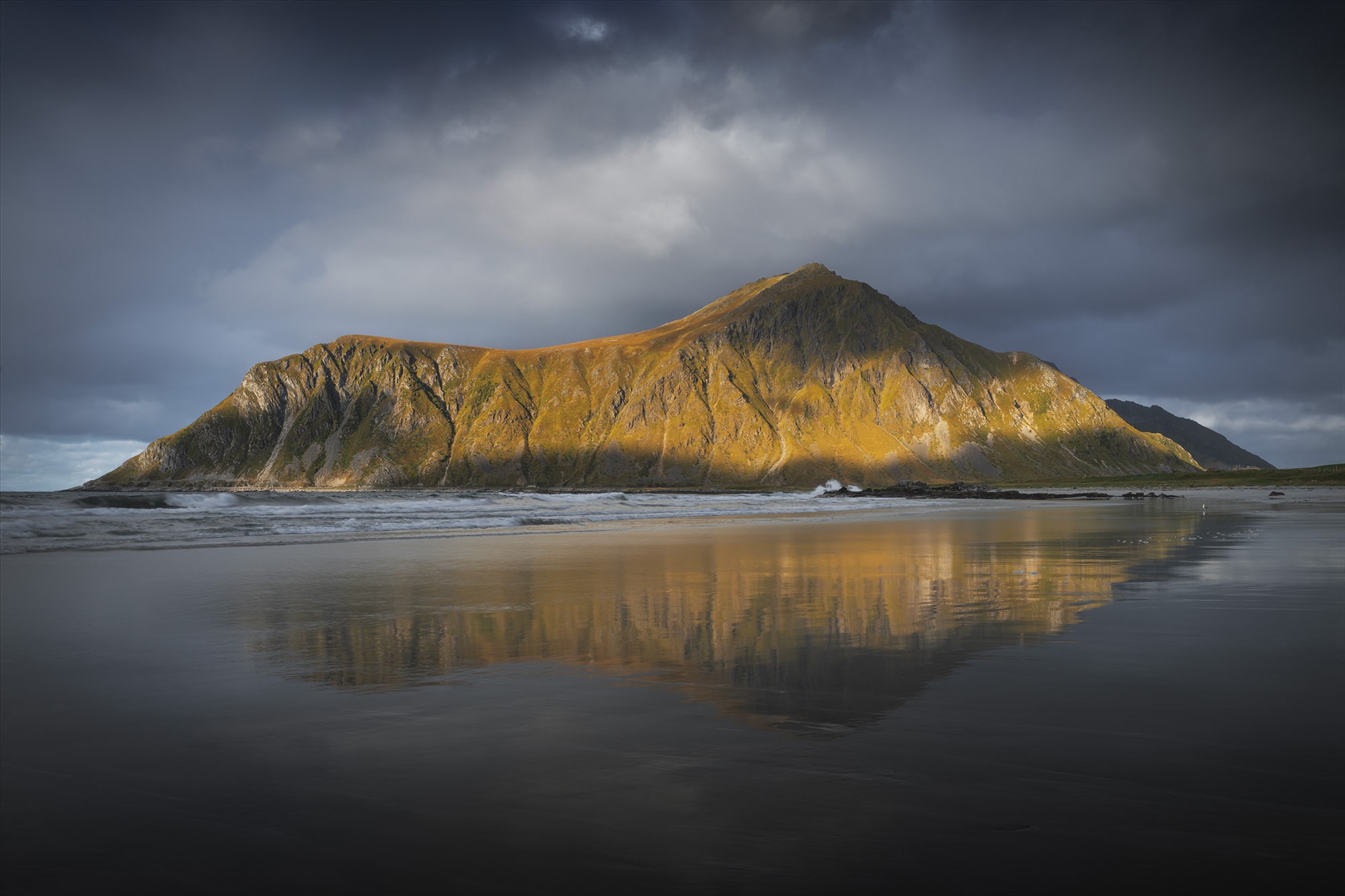 Golden sunlight hitting mountain at Skagsanden Beach, Lofoten Islands, Norway, with dramatic sky and reflection on wet sand during autumn.