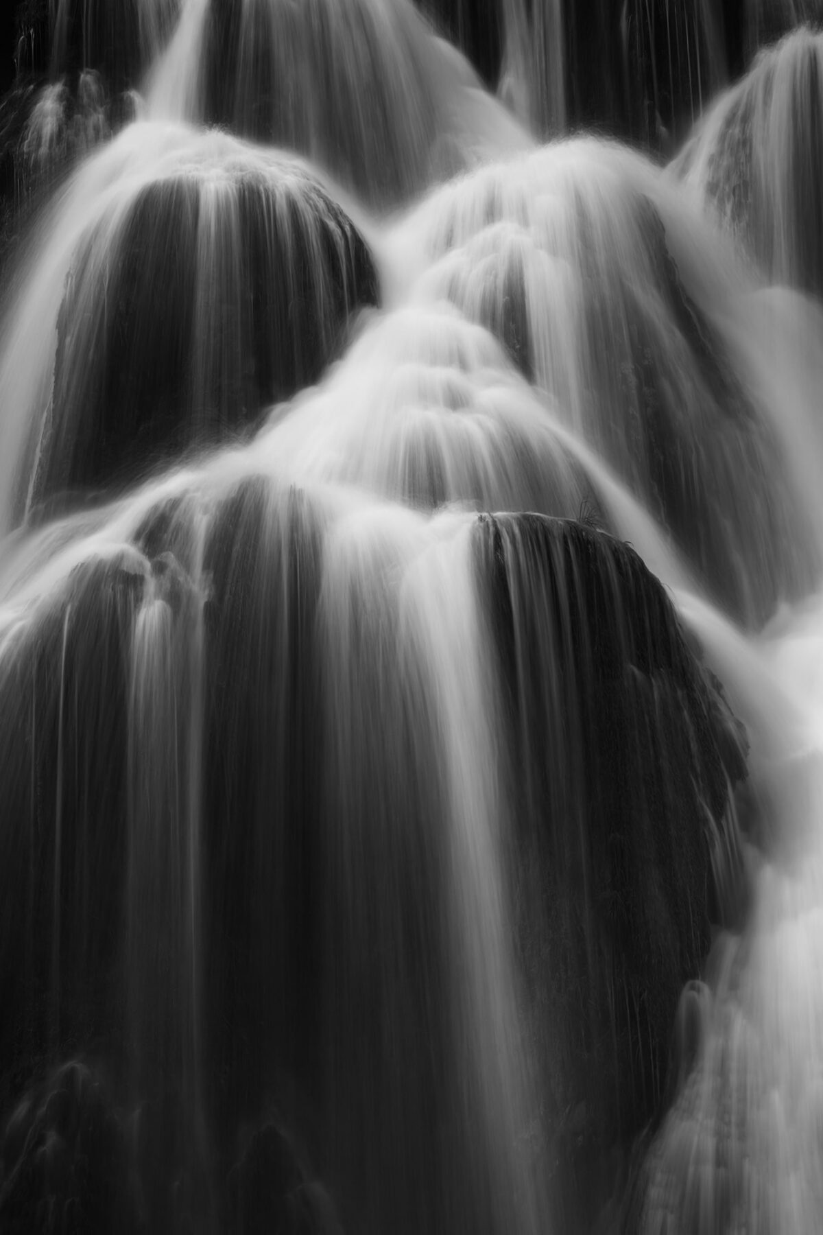 Black and white photograph of flowing water cascading over moss-covered rocks at Baume-les-Messieurs waterfall, Jura, France, captured with long exposure to emphasize movement and texture.