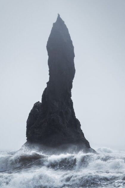 Photograph of a sea stack at Reynisfjara Beach in Iceland, with waves crashing below and a seagull standing still on top against a plain sky.