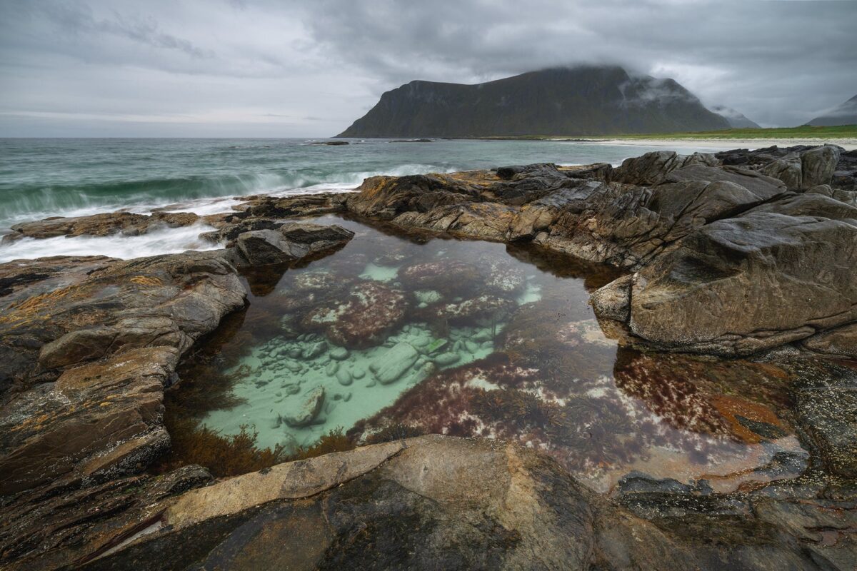 Turquoise tidal pool surrounded by coastal rocks at Skagsanden Beach in the Lofoten Islands, Norway, with moody clouds and mountain backdrop in autumn.
