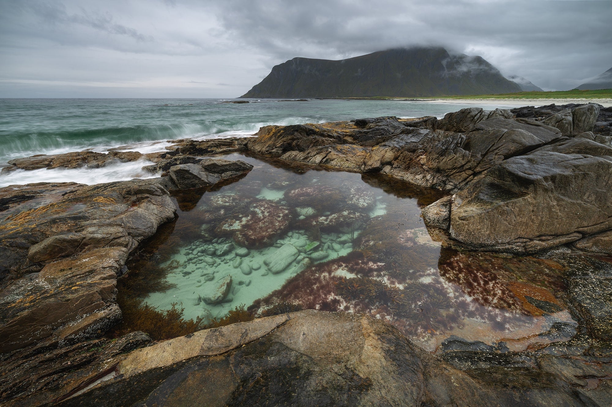 Turquoise tidal pool surrounded by coastal rocks at Skagsanden Beach in the Lofoten Islands, Norway, with moody clouds and mountain backdrop in autumn.