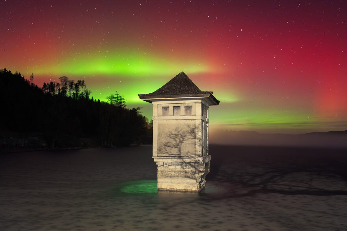 Landscape photography of the northern lights in Switzerland captured on January 19, 2026, at Lac de Bret in Lavaux, Canton of Vaud. Red and green aurora borealis glow above the lake, illuminating a small historic structure under a star-filled sky.