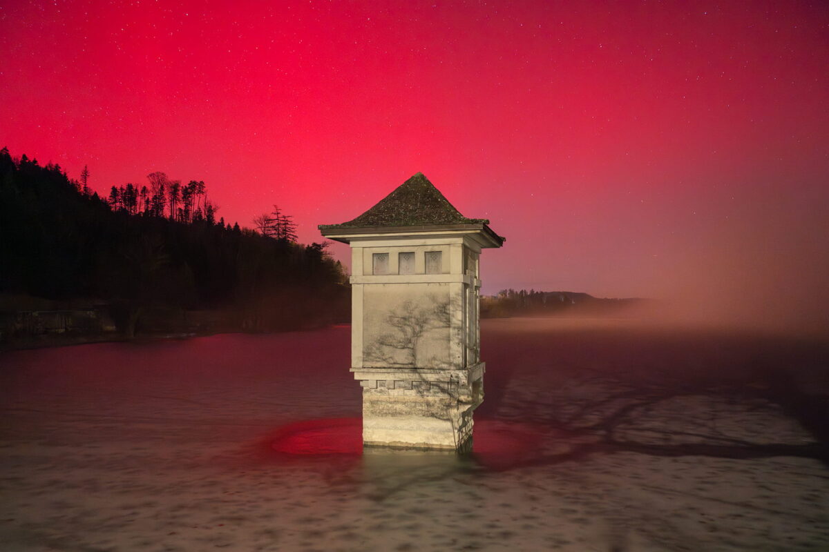 Landscape photography of a red aurora borealis with fog in Switzerland, captured on January 19, 2026, at Lac de Bret in Lavaux, Canton of Vaud. Crimson northern lights glow through mist above the lake, creating a dramatic and rare atmospheric scene.