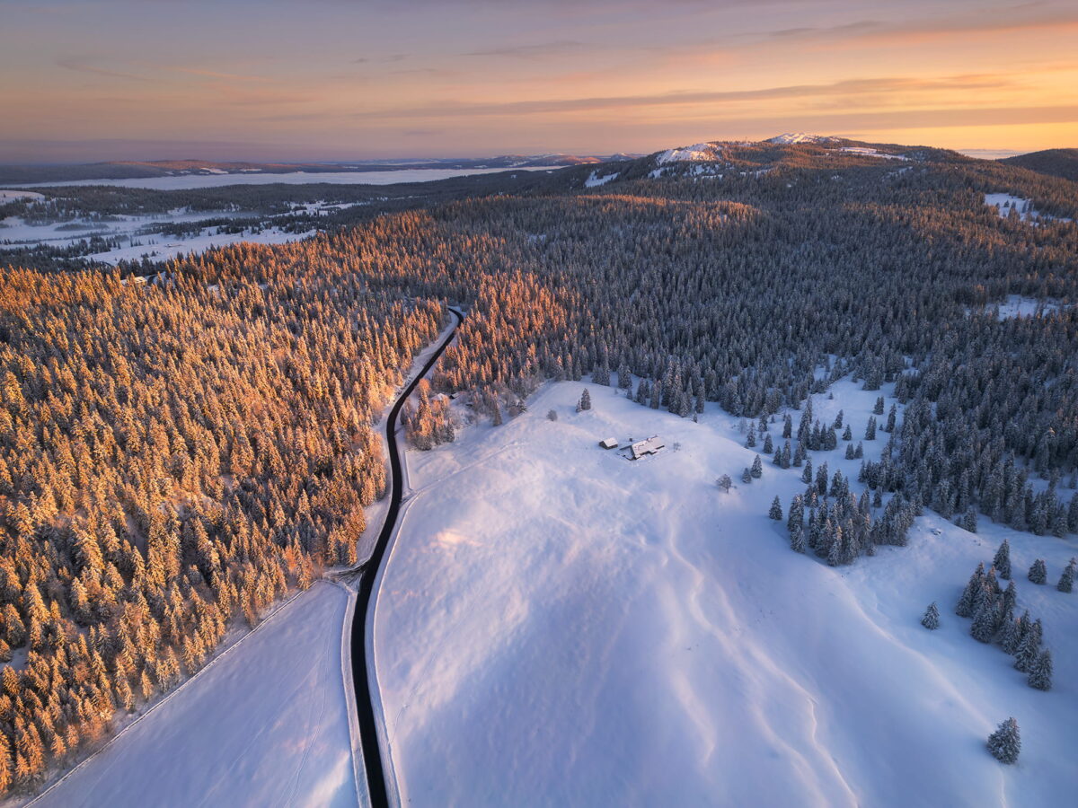 erial landscape photography of a snowy sunrise at Col du Marchairuz in Switzerland, captured by drone in the Parc du Jura Vaudois, with snow-covered forests glowing in early morning light.