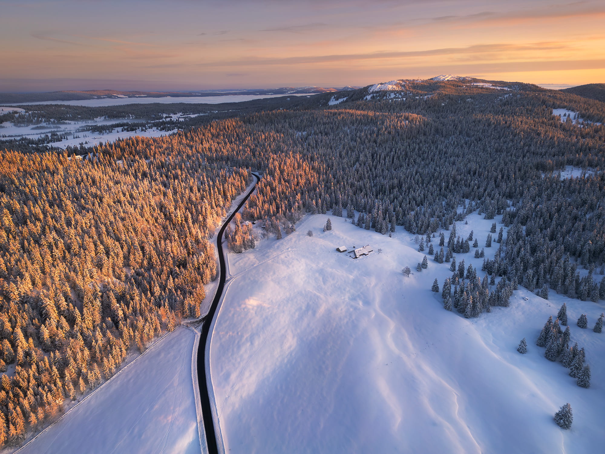 erial landscape photography of a snowy sunrise at Col du Marchairuz in Switzerland, captured by drone in the Parc du Jura Vaudois, with snow-covered forests glowing in early morning light.