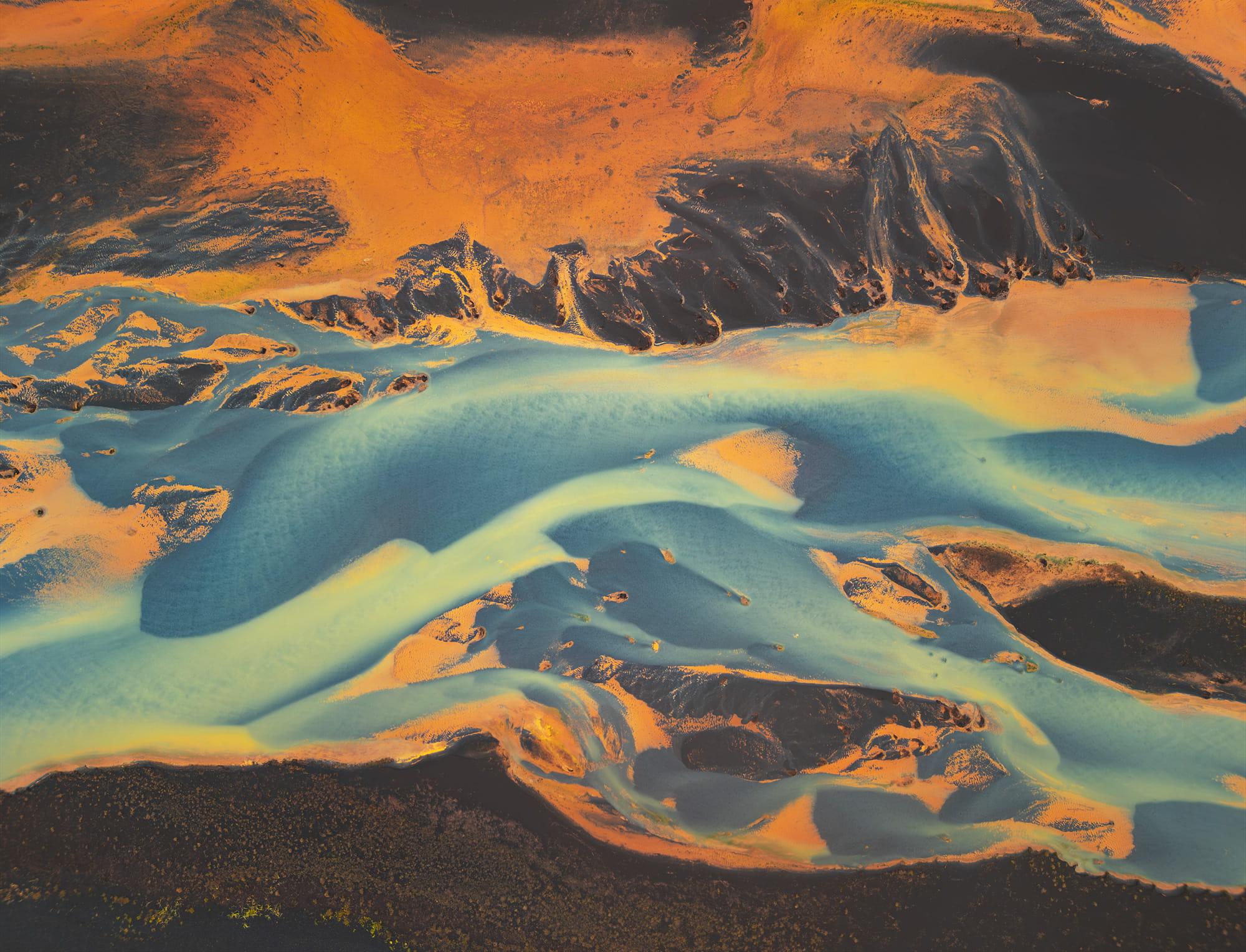 Drone landscape photography of braided rivers in southern Iceland, captured from an aerial view. Blue and yellow glacial waters flow through vivid orange volcanic ground, creating an abstract and graphic natural pattern.