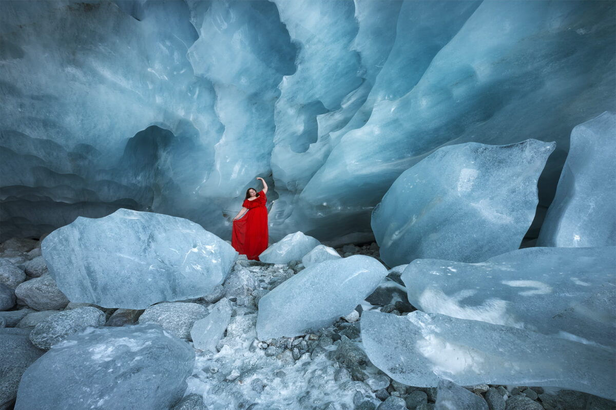 Self-portrait landscape photography in the Zinal ice cave, Valais (Wallis), Switzerland, featuring a woman in a red dress standing inside the blue glacial cave.